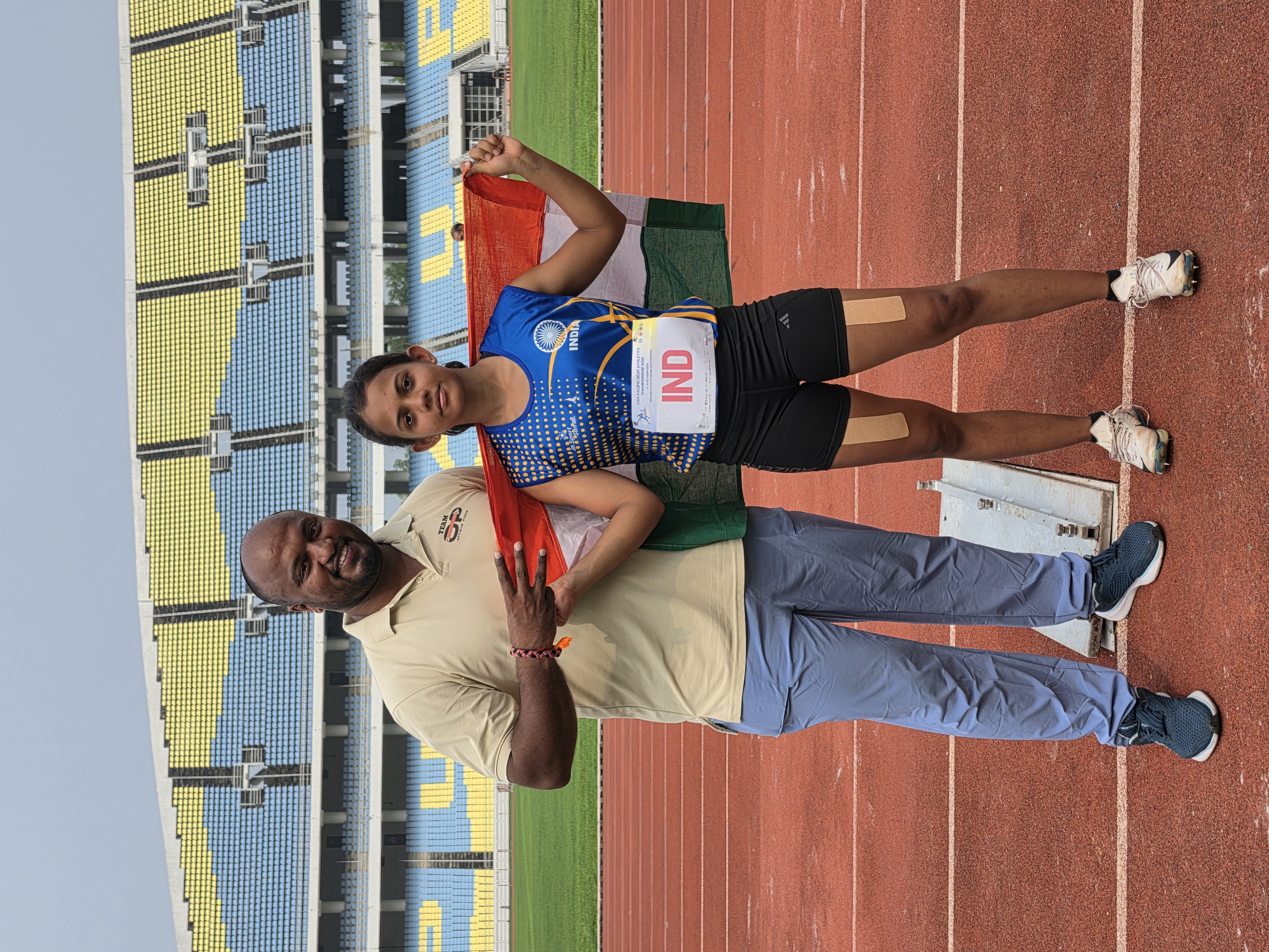 Rishitha in her India sprinting kit beside coach Radesh, holding the Indian flag at the stadium